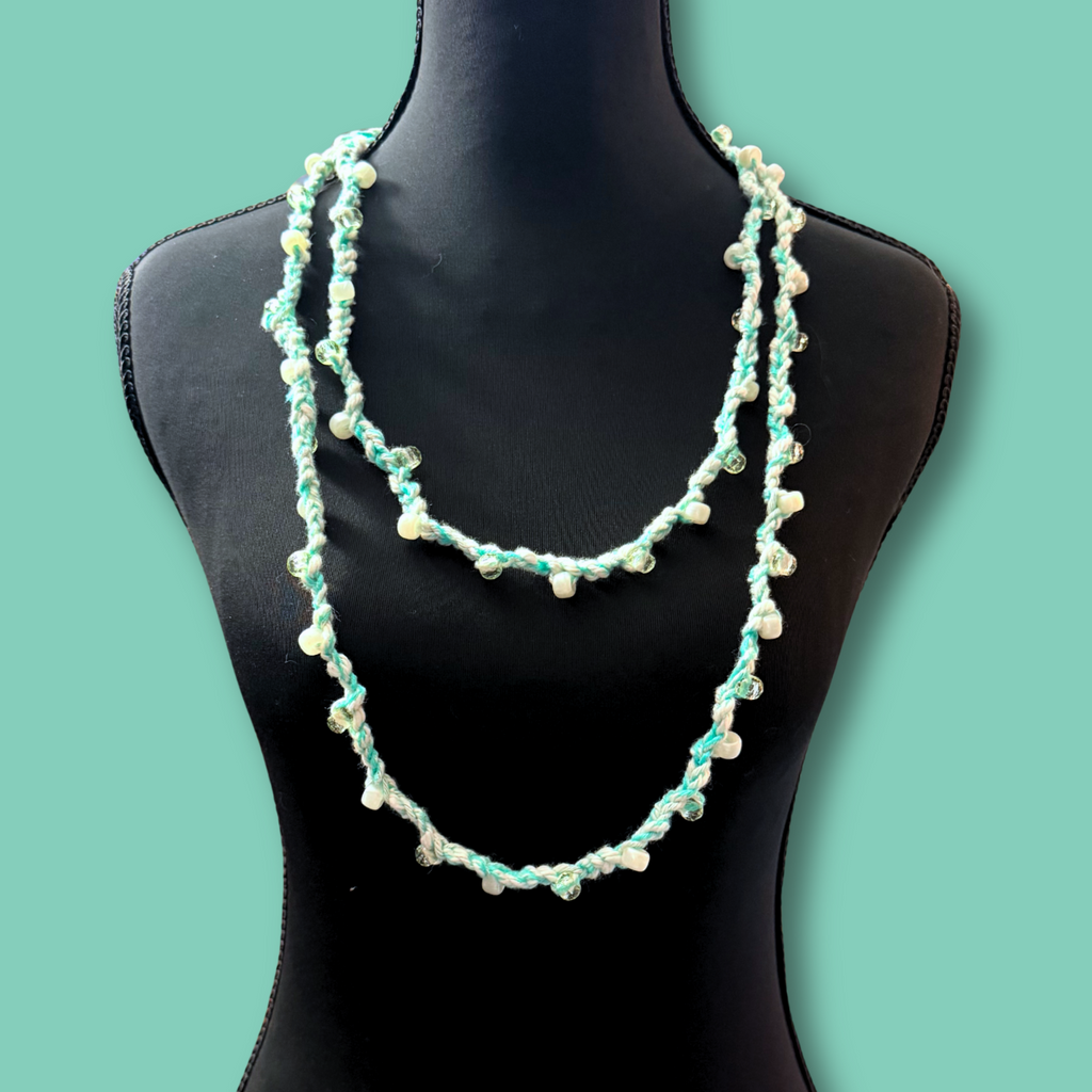 A two-strand beaded necklace displayed on a black mannequin torso against a solid mint green background. The necklace features light blue-green beads and small white pearl-like beads.