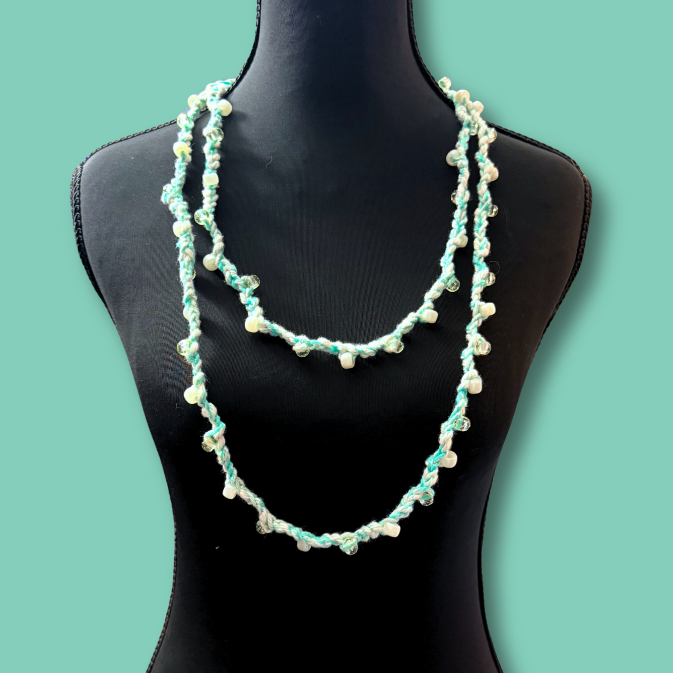 A two-strand beaded necklace displayed on a black mannequin torso against a solid mint green background. The necklace features light blue-green beads and small white pearl-like beads.