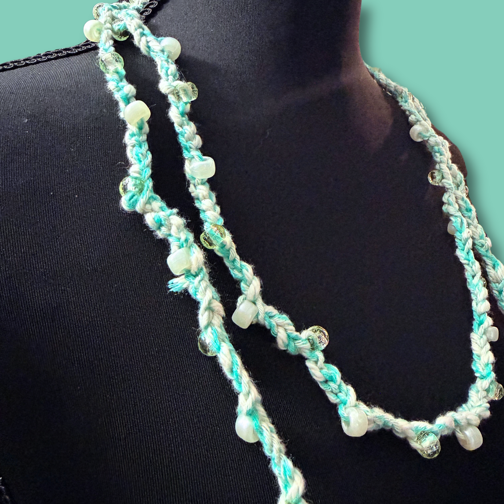 A long braided necklace in mint green and white yarn with small clear glass beads, displayed on a black mannequin bust against a solid teal background.